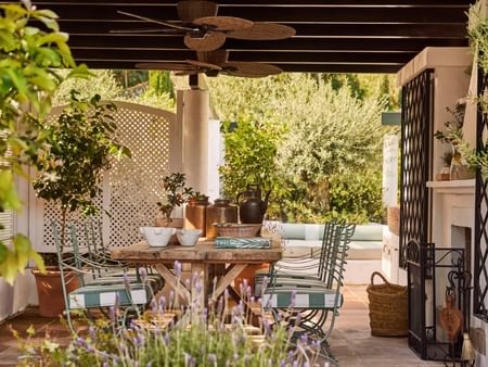 Patio con mesa de madera, sillas, ventiladores de techo, plantas y una chimenea decorativa.