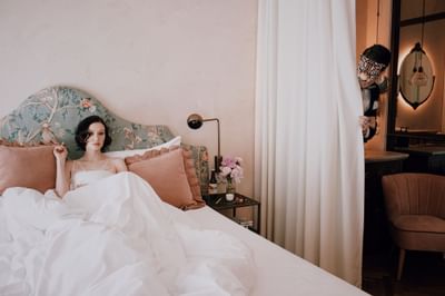 Lady resting in a plush pillow-filled bed while another peeks from behind the curtain in a cozy room at Hotel Motto Vienna
