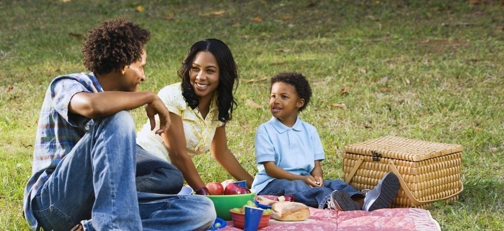 Family enjoying a picnic together on a grassy field with a picnic basket, fruit and snacks.