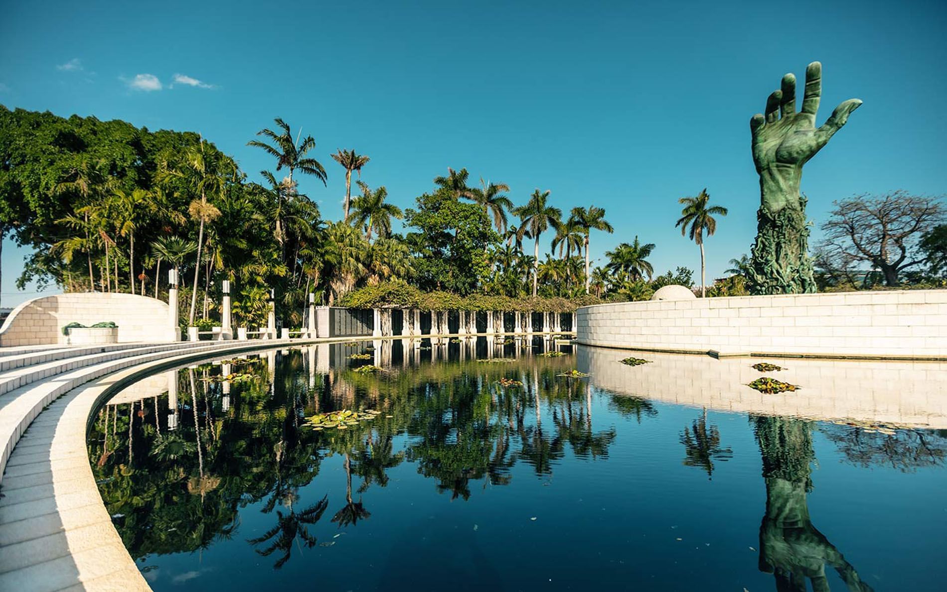 A large statue of a hand is seen above a pool with palm trees in the background.