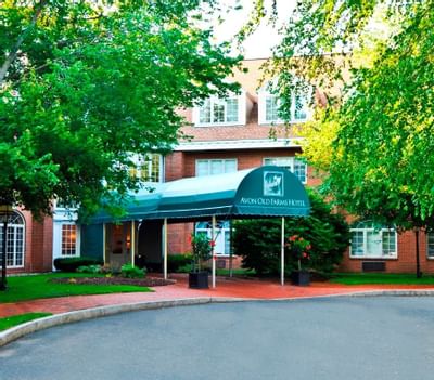 Front entrance driveway with green awning over brick building facade between green lawn and trees at Avon Old Farms Hotel