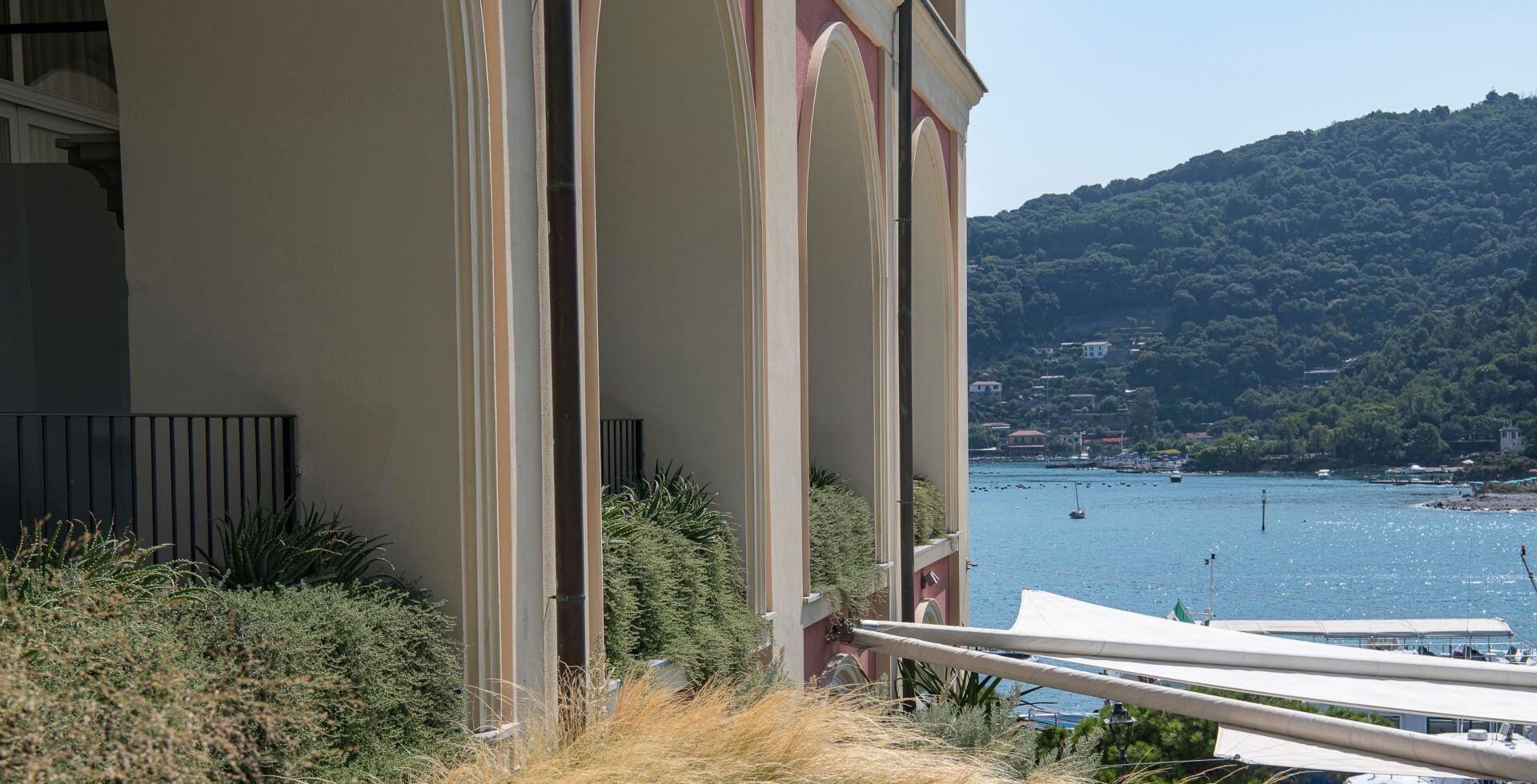 Vue depuis un balcon sur la mer, une colline boisée et un bâtiment avec des arches.
