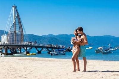 Couple walking on sandy beach with modern pier and mountain backdrop at Playa Los Arcos Puerto Vallarta
