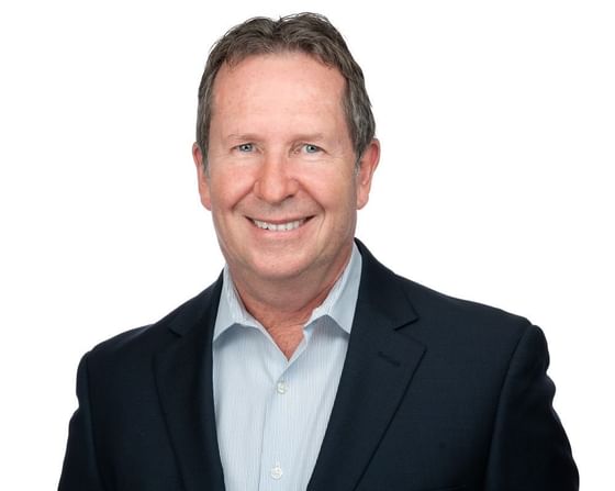 A smiling man in a blue shirt and navy blazer stands against a plain white background.