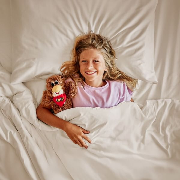 Girl lying in bed with white sheets, holding a stuffed animal, and smiling at the camera.