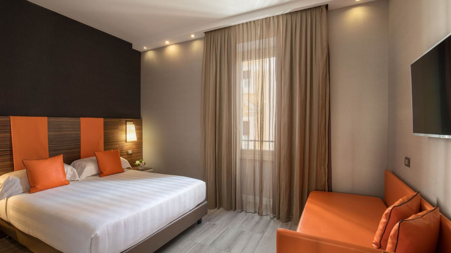 Deluxe Room featuring a double bed with orange pillows, a small sofa, a TV, and curtains at The Republic Hotel