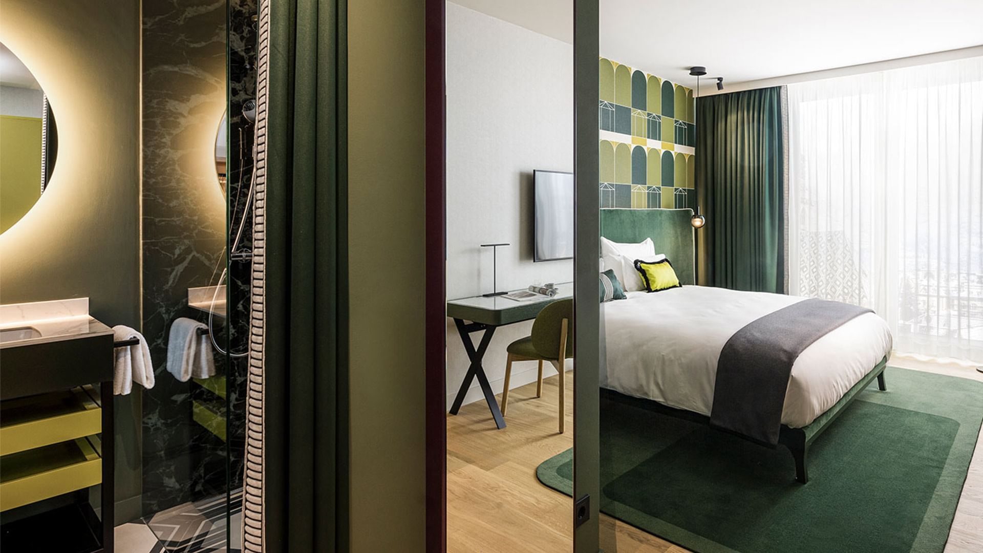 Zimmer mit Bad, Schreibtisch, Bett, Sessel, Teppich und Balkon mit Blick auf die Stadt.
