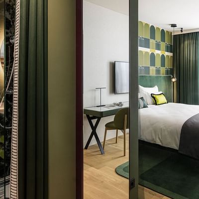 Zimmer mit Bad, Schreibtisch, Bett, Sessel, Teppich und Balkon mit Blick auf die Stadt.