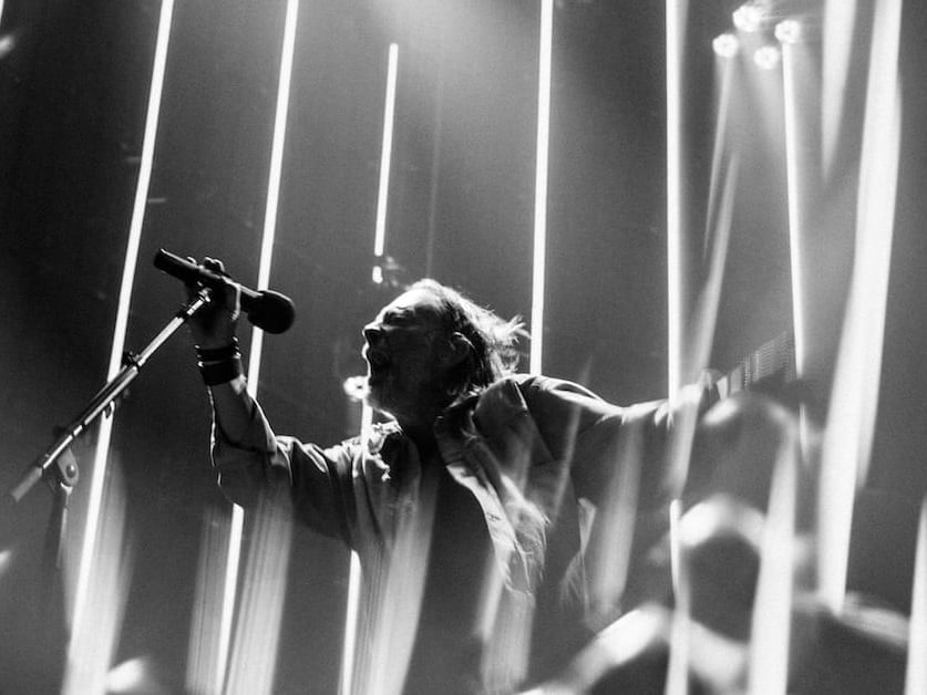 Black and white photo of a performer singing on stage with dramatic vertical light beams for Radiohead 2027