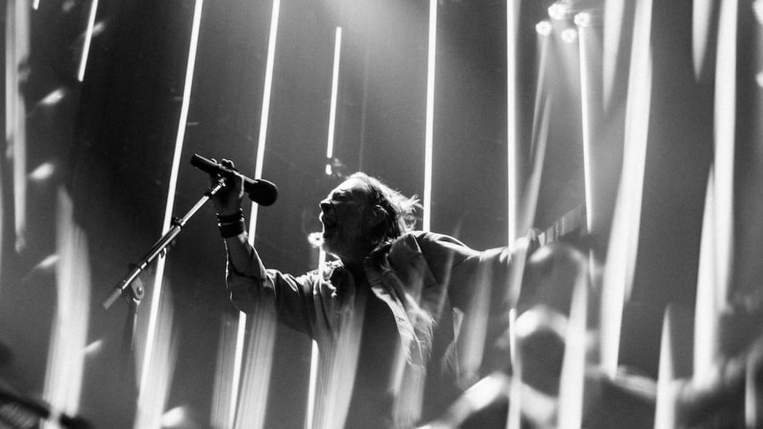 Black and white photo of a performer singing on stage with dramatic vertical light beams for Radiohead 2027