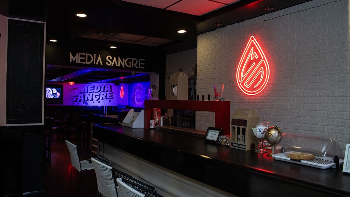 Red neon teardrop logo on a brick wall beside a dark bar counter with seating in Media Sangre at Araiza Calafia
