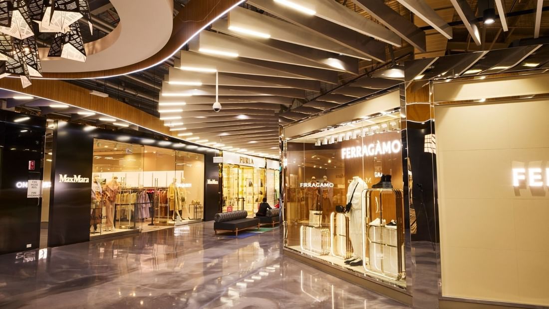 Modern shopping mall interior with luxury boutiques, polished marble floors, and contemporary wooden ceiling with integrated lighting.