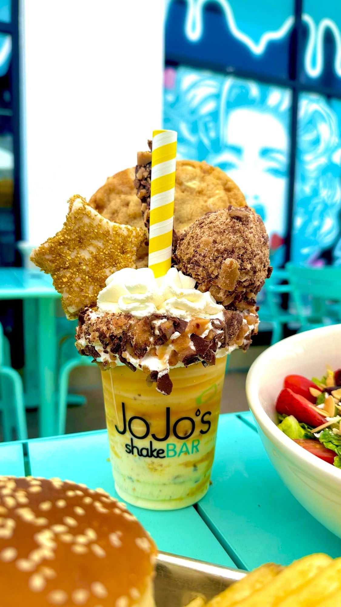 A milkshake topped with a cookie, a gold star marshmallow, whipped cream, and candy in a cup that reads JoJo's ShakeBAR sits on a blue table beside a burger and a salad.