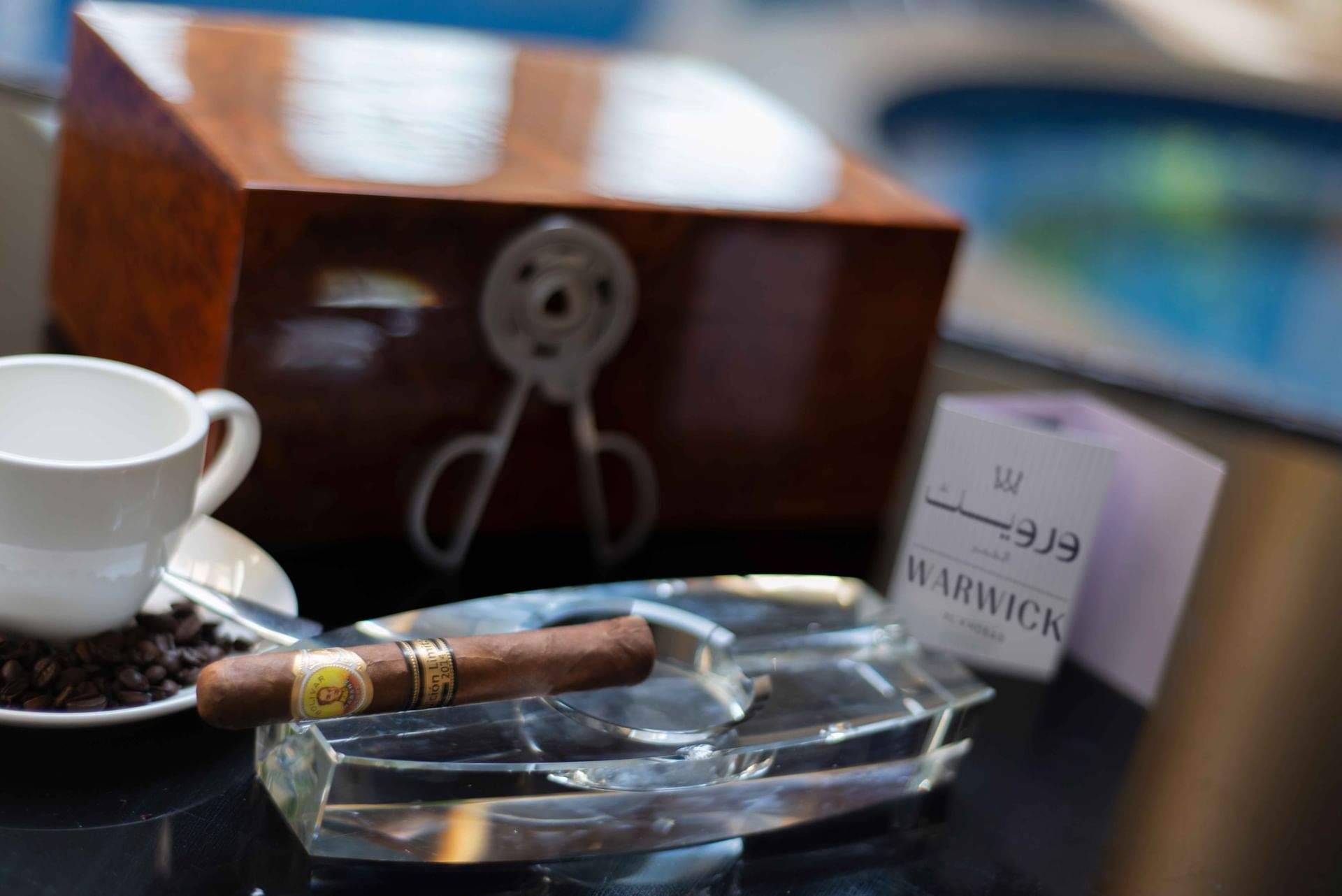 Single cigar rests in a clear crystal ashtray on a dark table next to a coffee cup in Cigar Lounge at Warwick Al Khobar