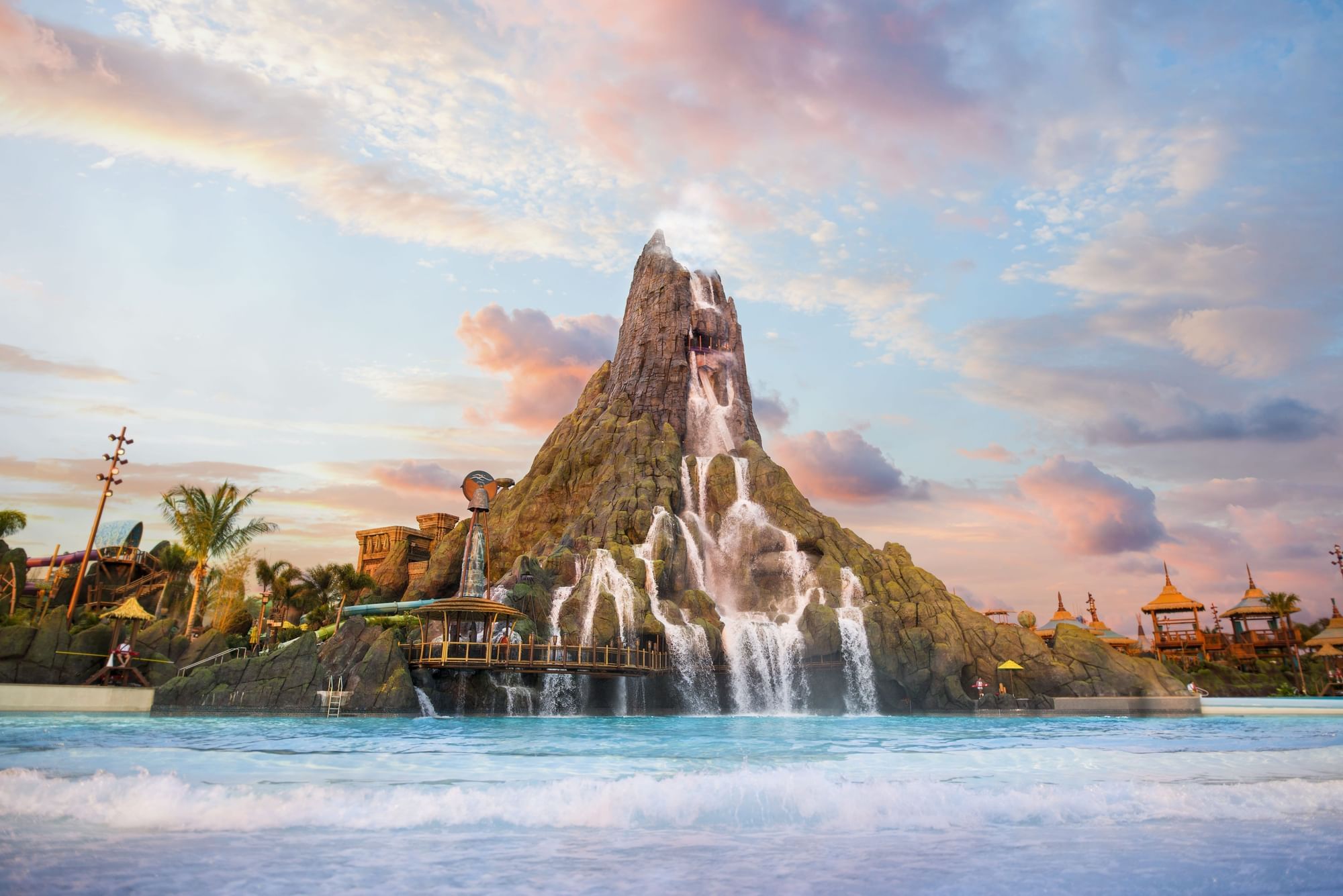 A tall, rocky volcano with a waterfall cascading down the side and falling into a surrounding blue wave pool.