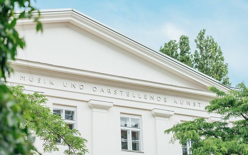 Klassizistische Fassade der Universität für Musik und darstellende Kunst Wien mit Schriftzug am Giebel, umrahmt von Bäumen.