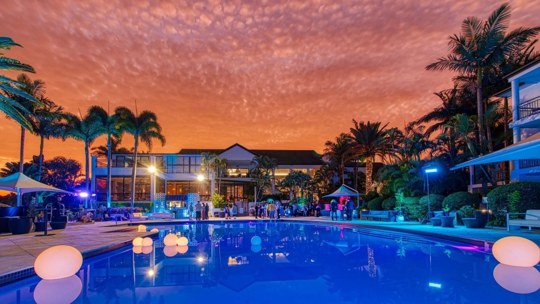 Illuminated resort pool at dusk with colorful sky, people, and palm trees.