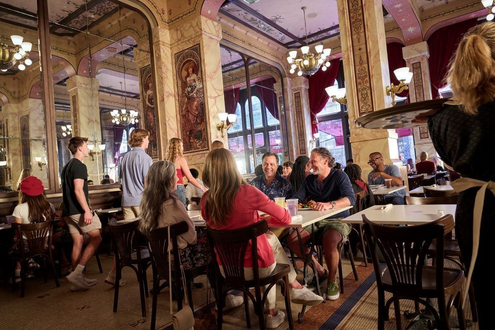 Guests dine at tables in a Parisian-inspired cafe with marble walls and ornate chandeliers. 