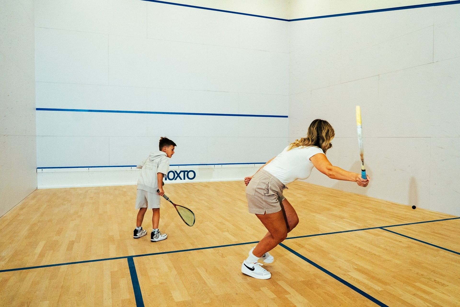 Woman and boy playing squash inside a wood floor court with high white walls and blue lines at Hotel X Toronto