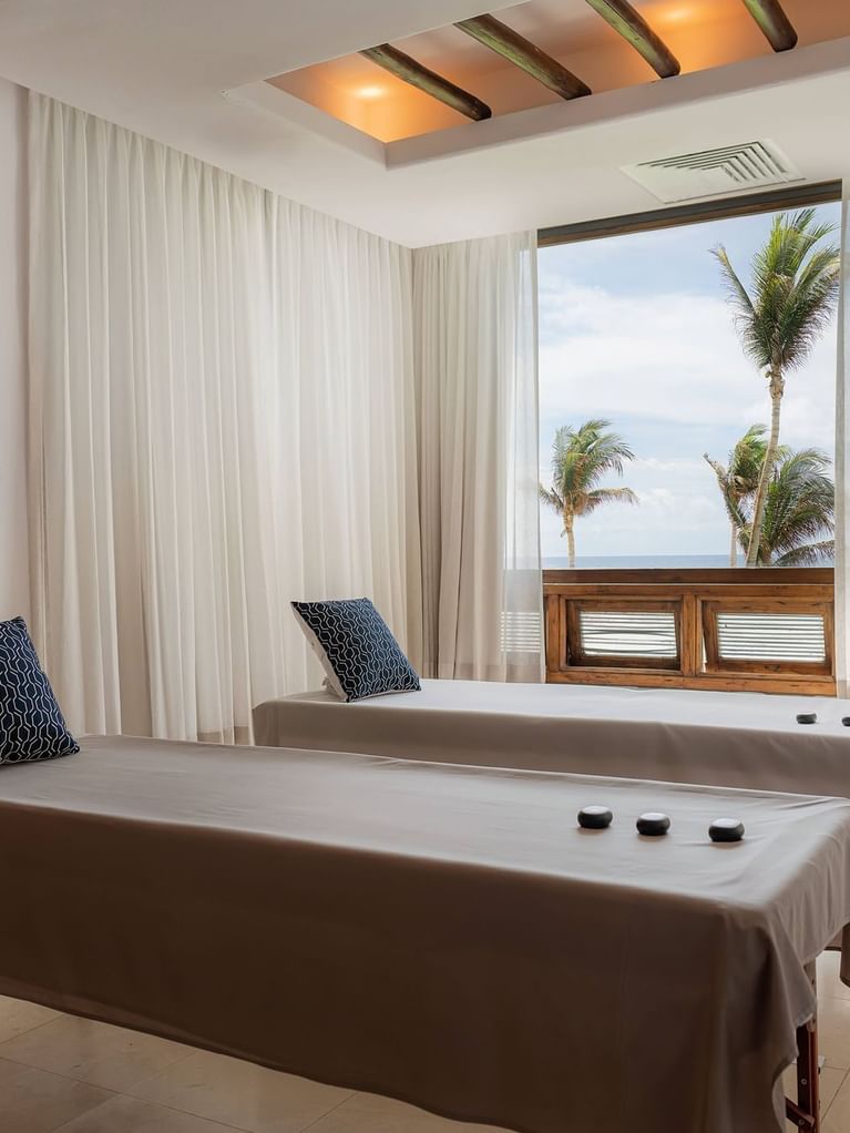 Massage treatment room with ocean view, bed with stones, palm trees visible outside.
