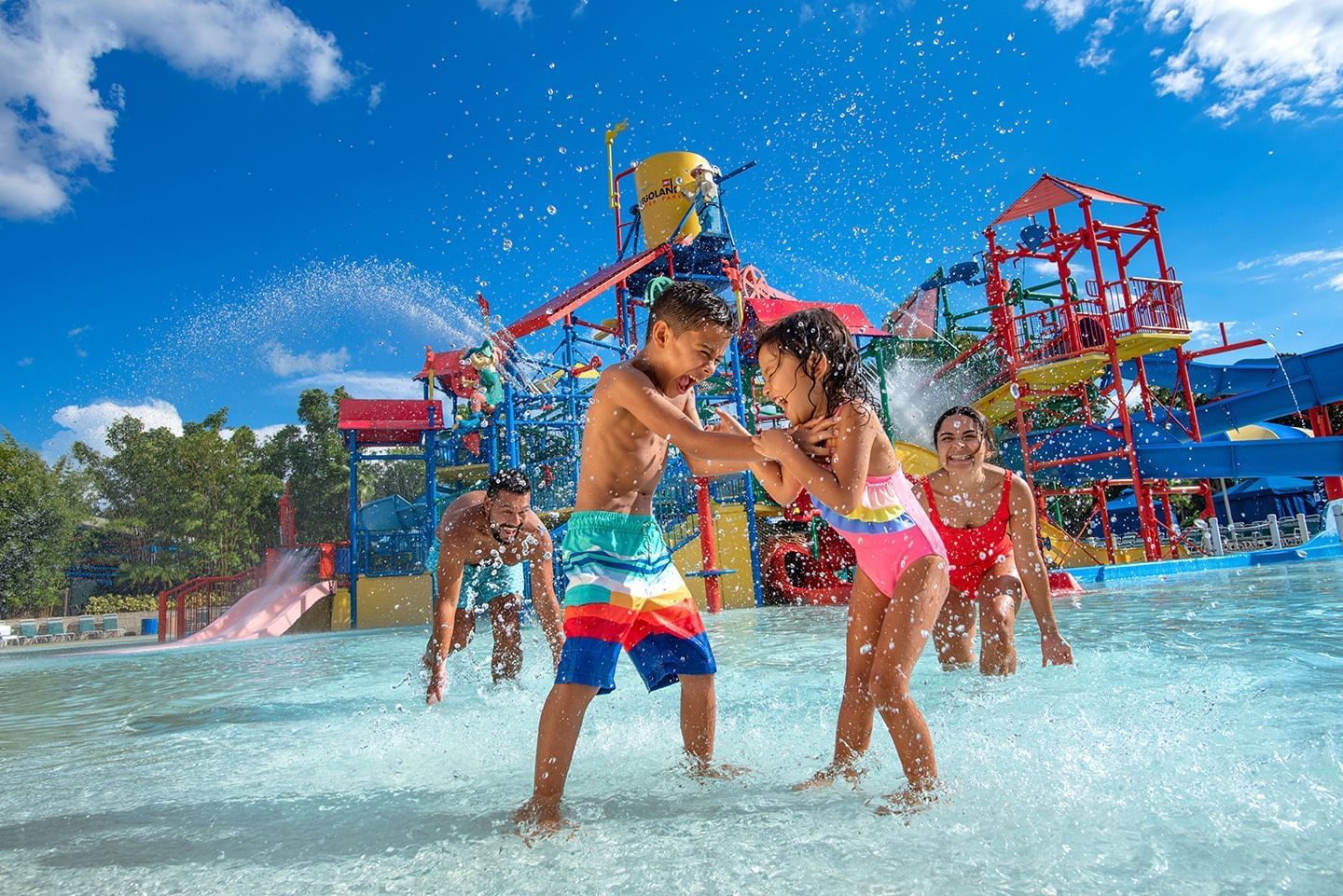 A boy and a girl in bathing suits splash one another in a large splash pad with their parents and a large play structure behind them.