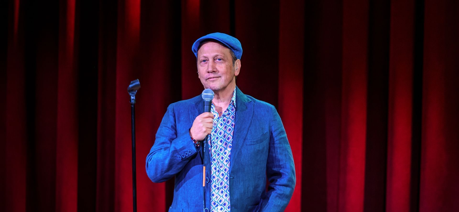 Actor and comedian Rob Schneider wearing blue cap holding microphone on stage with red curtain at The Brook