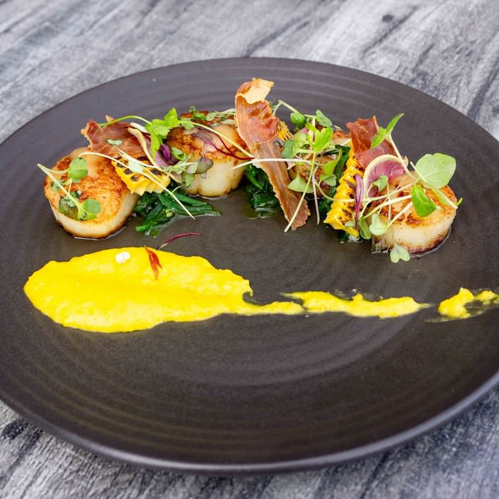 A black plate with scallops, bacon, greens, and yellow sauce on a wooden surface.
