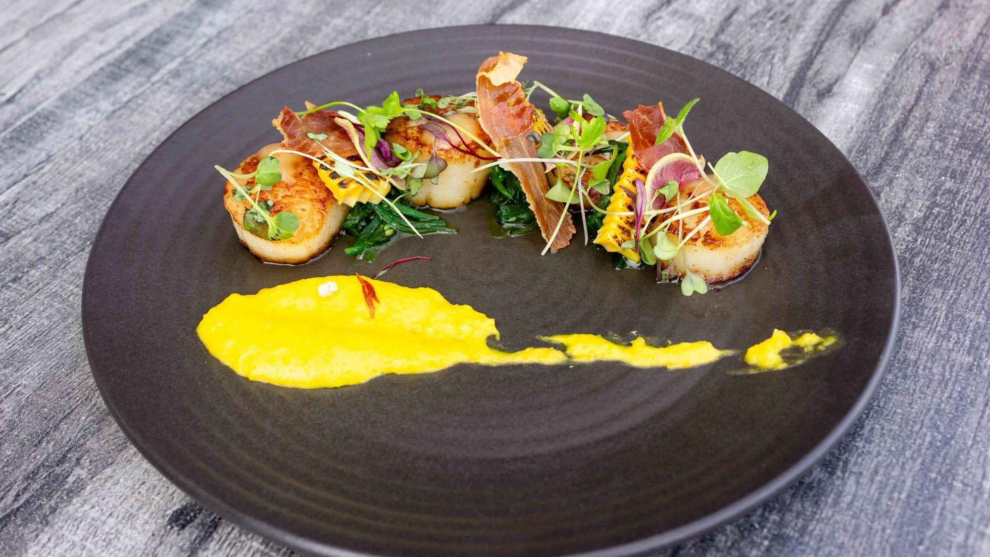A black plate with scallops, bacon, greens, and yellow sauce on a wooden surface.