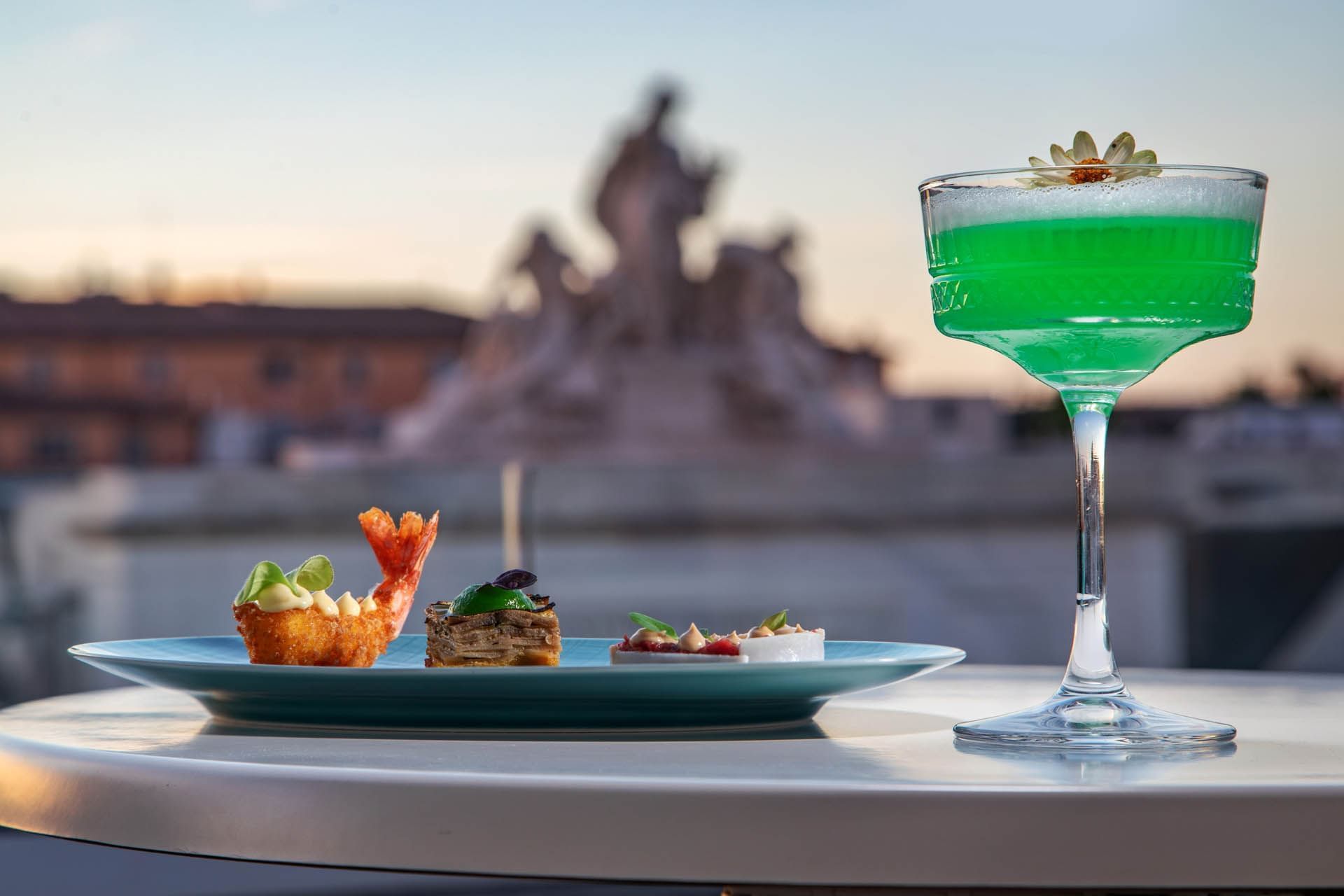 Green cocktail garnished with a flower beside an elegant plate of gourmet appetizers served at The Glam Hotel