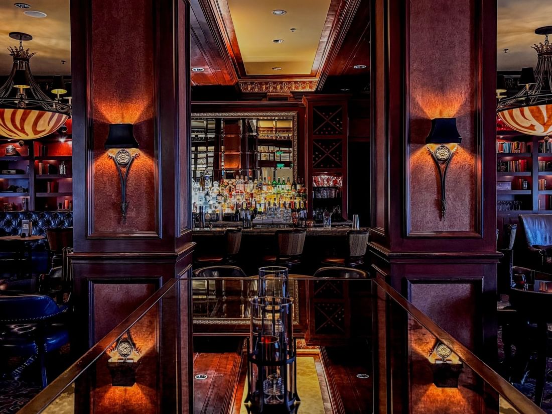 Polished wood table by bar stools under warm sconces in The Library at Warwick Melrose Dallas