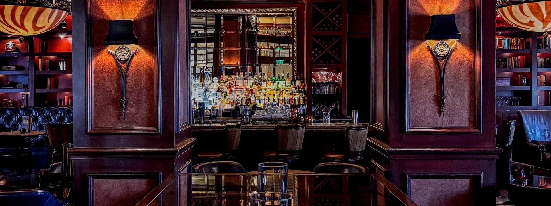 Polished wood table by bar stools under warm sconces in The Library at Warwick Melrose Dallas