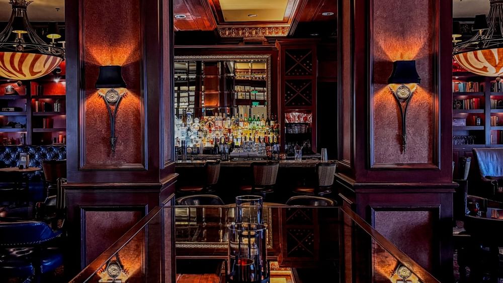 Polished wood table by bar stools under warm sconces in The Library at Warwick Melrose Dallas
