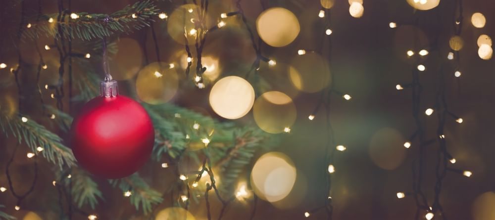 A close-up of a red ornament hanging on a Christmas tree branch surrounded by warm, fuzzy Christmas lights. 