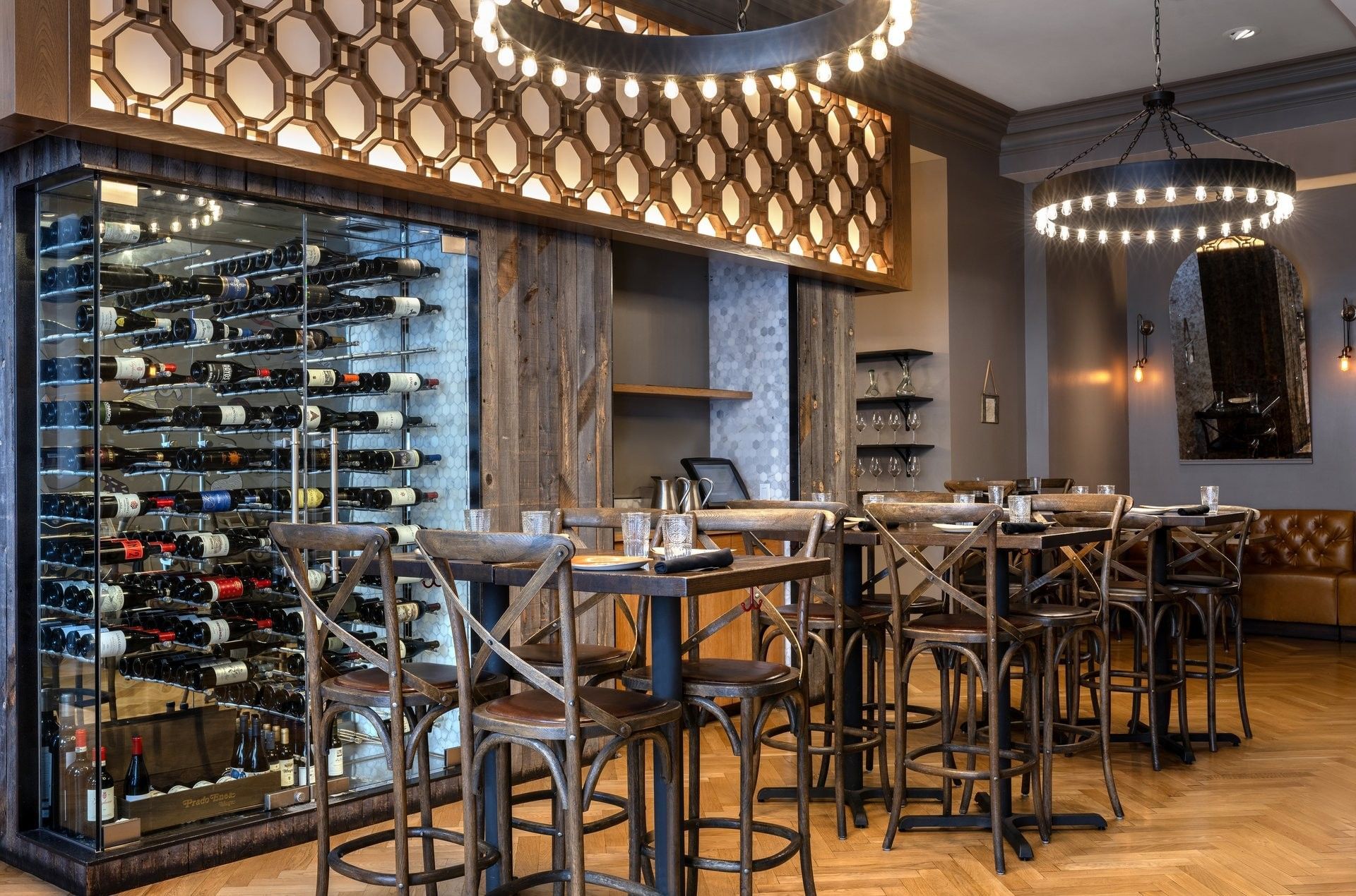 Wine bottles with a glass cellar by wooden tables and chairs under ring lights in BOTA at Warwick San Francisco