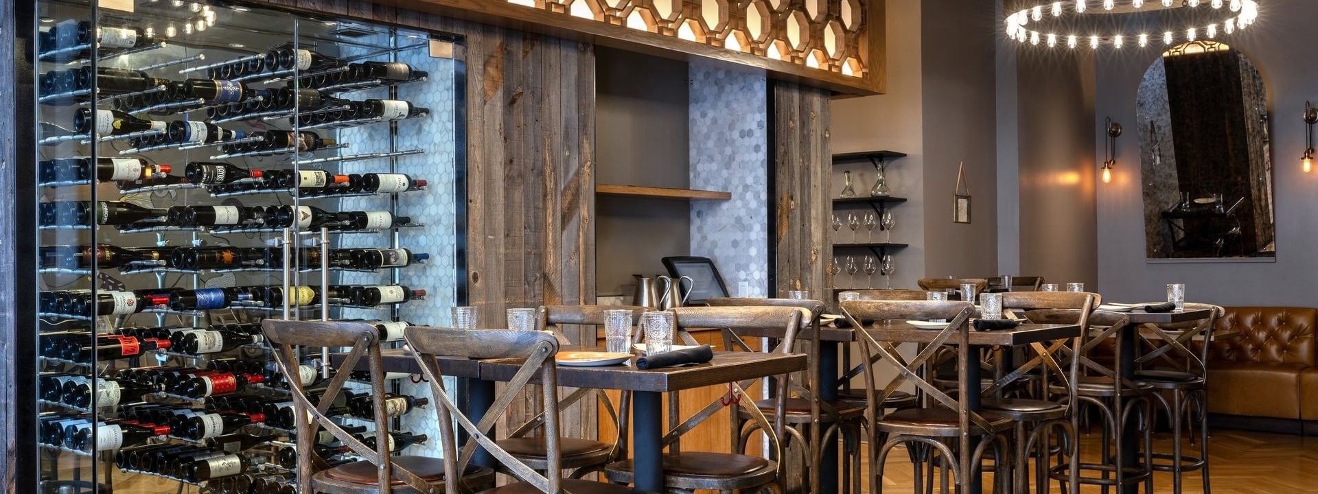 Wine bottles with a glass cellar by wooden tables and chairs under ring lights in BOTA at Warwick San Francisco