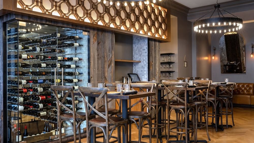 Wine bottles with a glass cellar by wooden tables and chairs under ring lights in BOTA at Warwick San Francisco