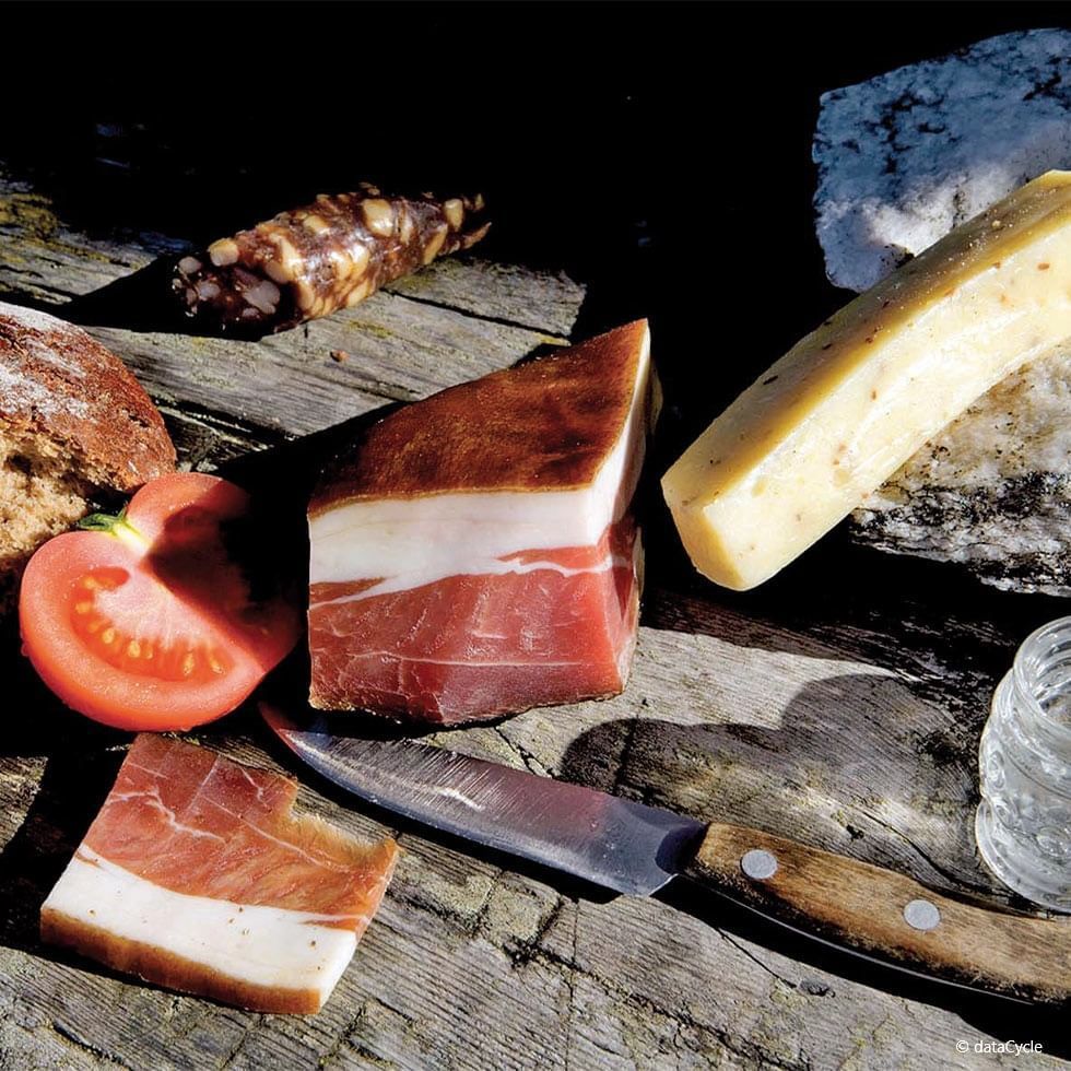 Coltello, prosciutto, formaggio, pane, pomodori e un bicchiere d'acqua su un tavolo in legno.