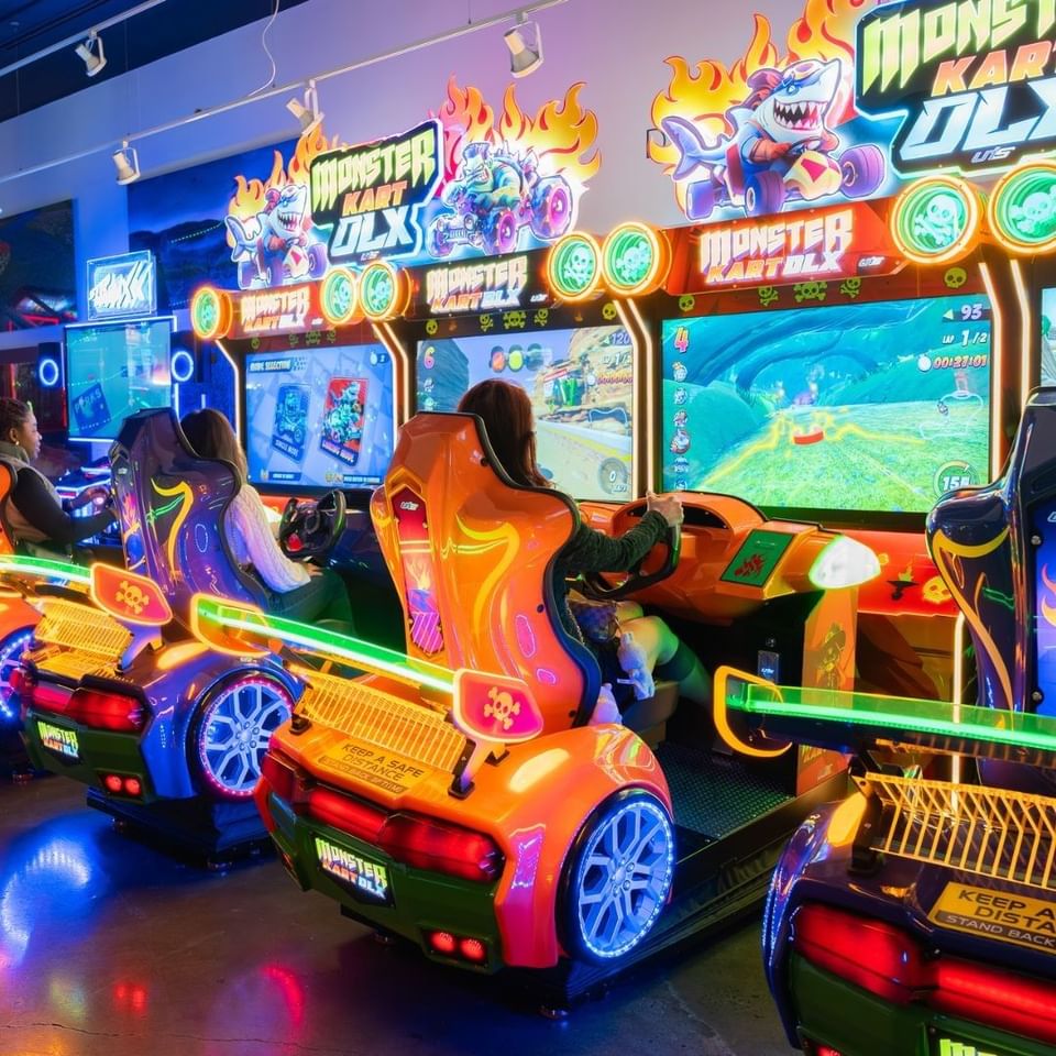 Guests seated in orange Monster Kart DLX arcade racers, under neon flame graphics and screens at Hotel X Toronto