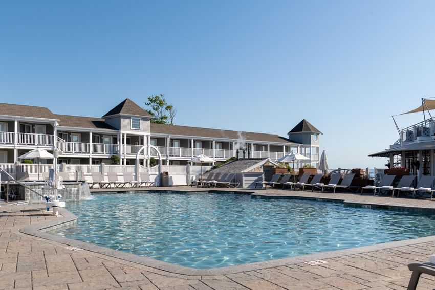 A large outdoor pool with lounge chairs and umbrellas in front of a multi-story hotel building with balconies.