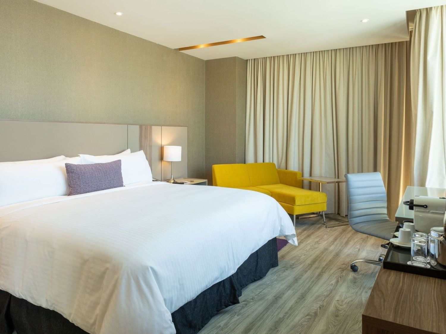 King Junior Suite with white bedding, yellow sofa, work desk, and floor-to-ceiling curtains at Camino Real Fashion Drive.