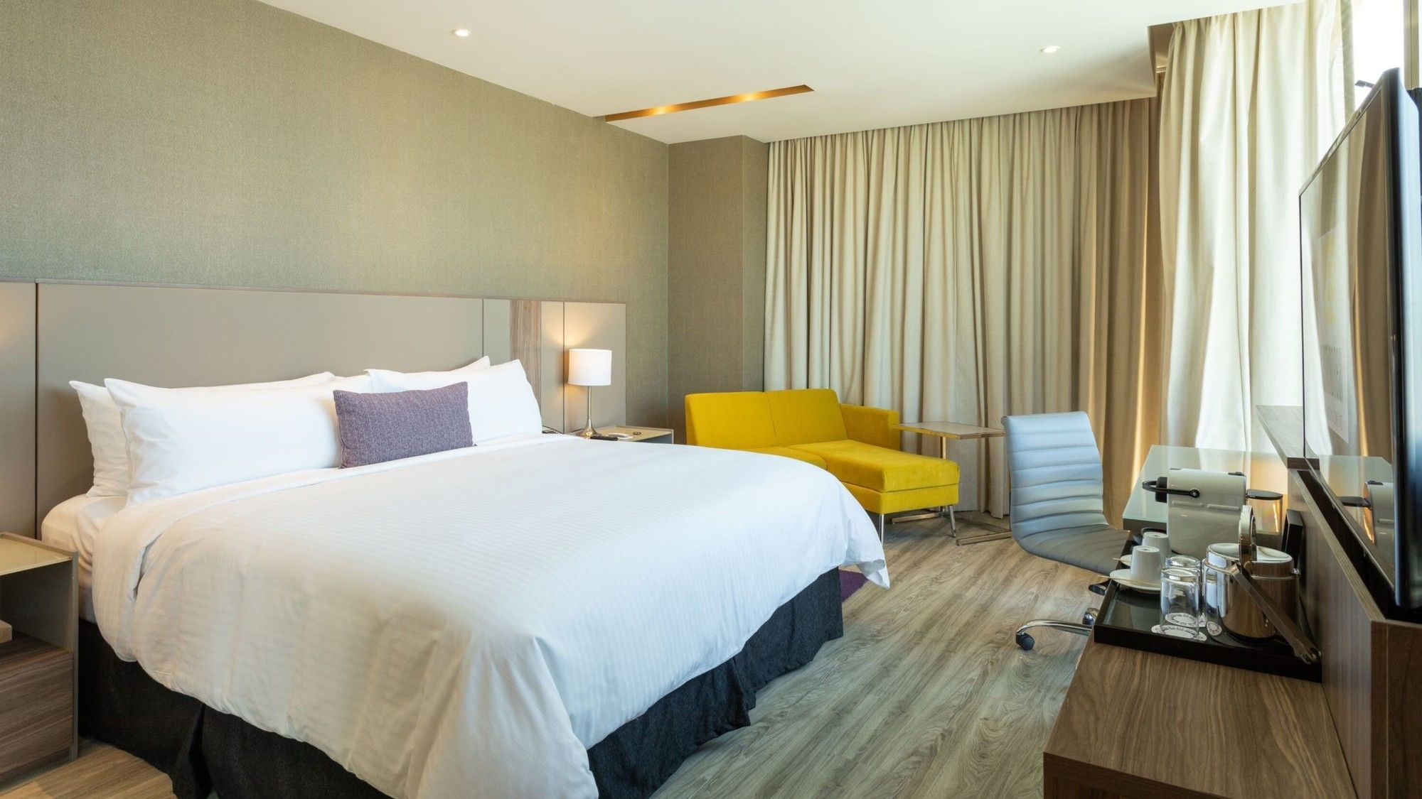 King Junior Suite with white bedding, yellow sofa, work desk, and floor-to-ceiling curtains at Camino Real Fashion Drive.
