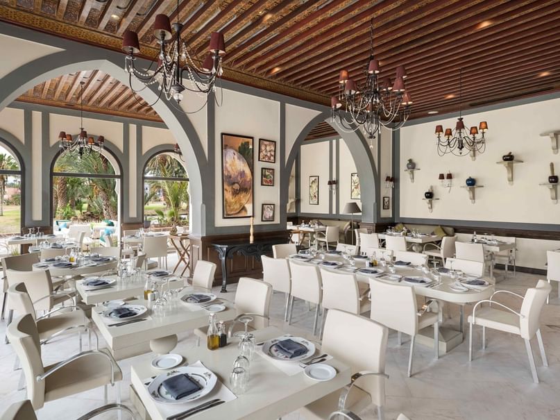 An elegant dining room with white tables and chairs, chandeliers, and large arched windows overlooking greenery.