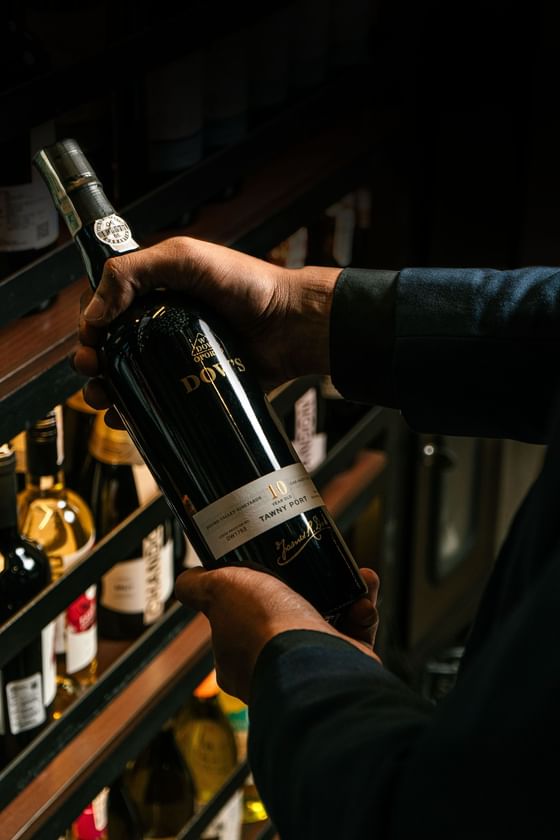 Close-up of hands holding a bottle of Dow's 10 Year Old Tawny Port wine in a dark cellar setting at Vasa Hotel Surabaya