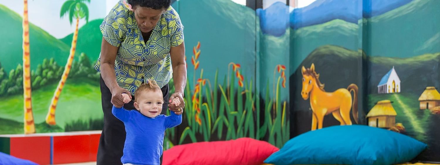 Nanny helping a toddler walk by colorful pillows under a mural in the kids club at Warwick Fiji Resort and Spa