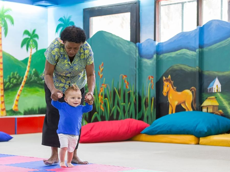 Nanny helping a toddler walk by colorful pillows under a mural in the kids club at Warwick Fiji Resort and Spa