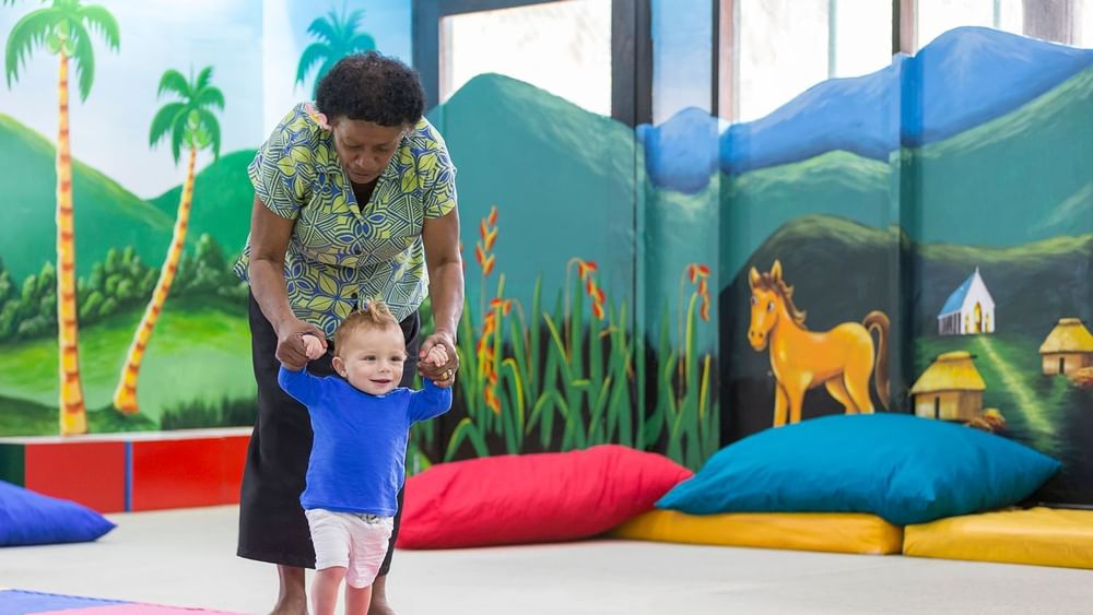 Nanny helping a toddler walk by colorful pillows under a mural in the kids club at Warwick Fiji Resort and Spa
