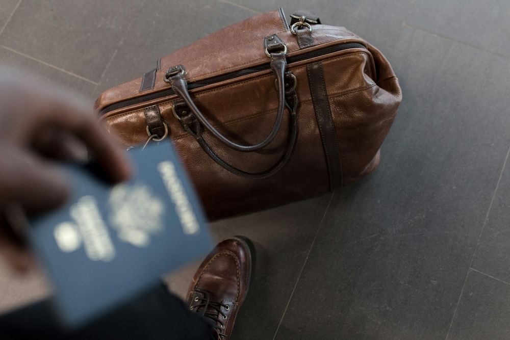 A view from above of a leather travel bag with a man’s shoe in frame, and a hand holding a passport out of focus in the foreground. 