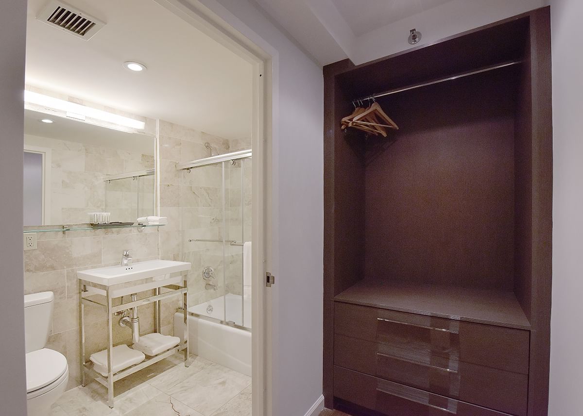 A bathroom with a shower, sink, and toilet, and a closet with drawers and a hanging rod.