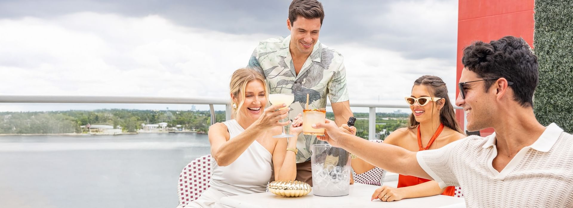 A group of four young adults enjoying drinks at an outdoor bar with a body of water in the background.