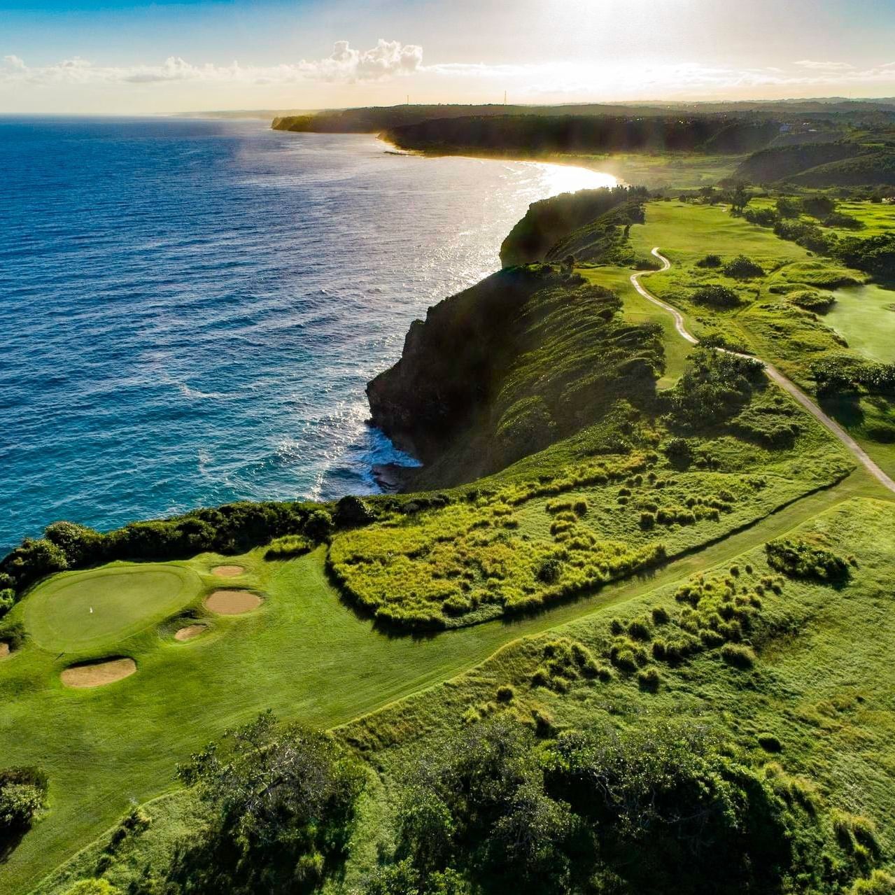 Aerial view of a scenic coastal golf course with lush green fairways, sand bunkers, and dramatic cliffs overlooking blue ocean waters.